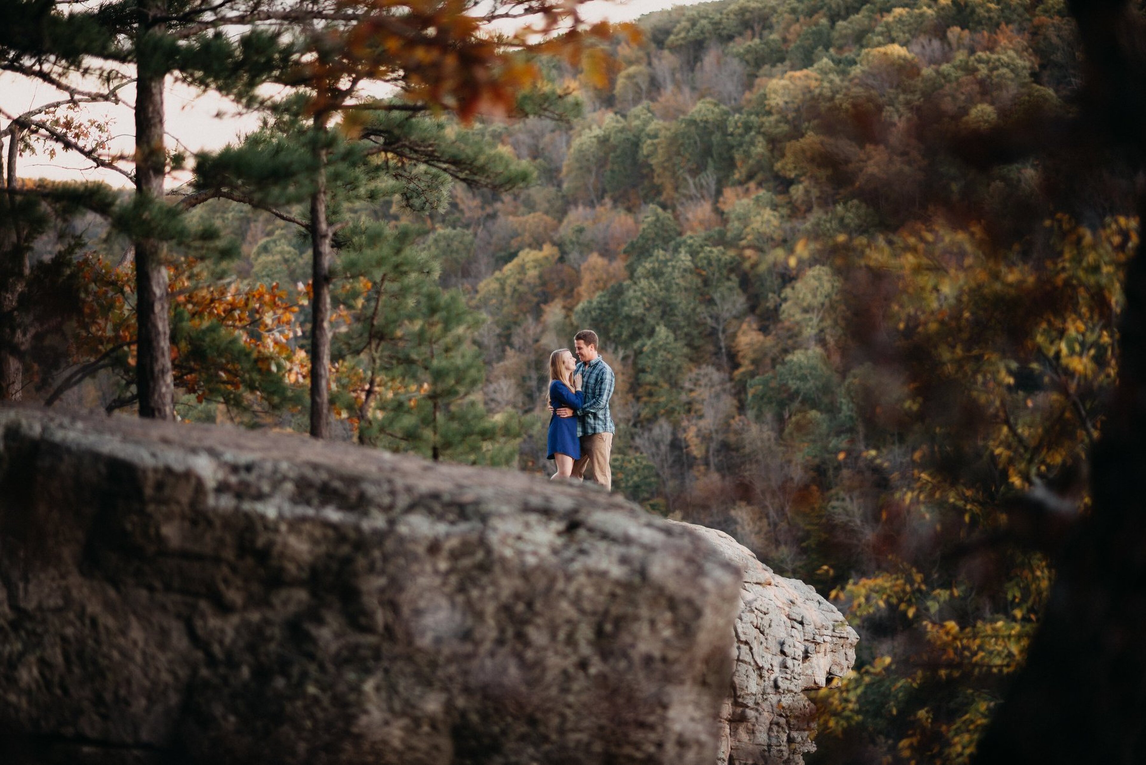 Couple standing together on a cliffside forest overlook with autumn trees and soft cinematic framing