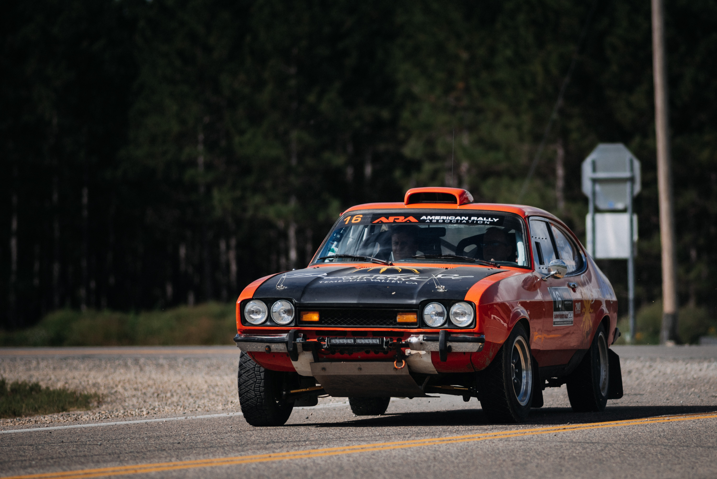Vintage orange Ford Escort rally car driving toward the camera on a paved road bordered by dark trees.