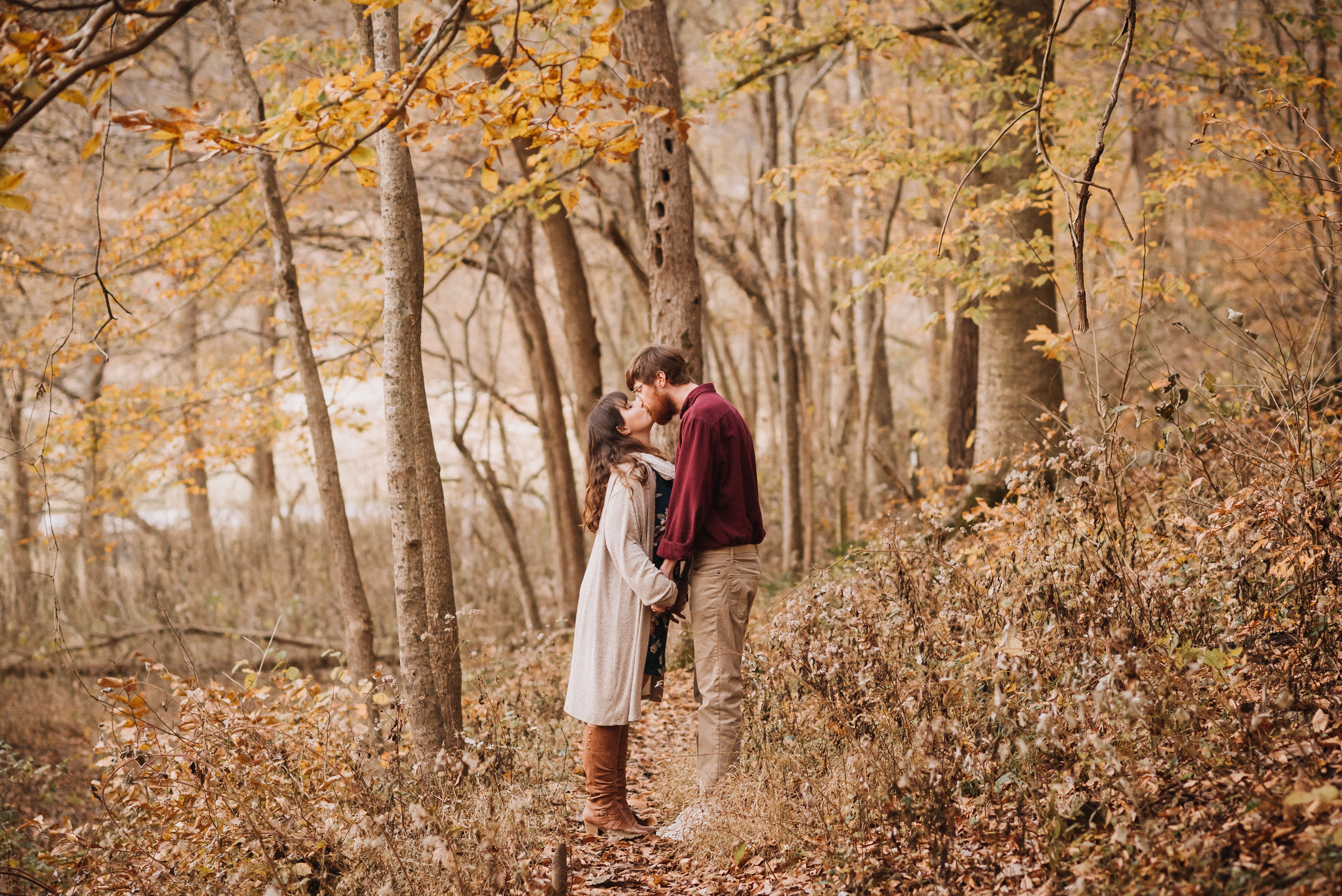 Matt and Carmen kissing on a leaf-covered forest trail in warm autumn light at Steel Creek