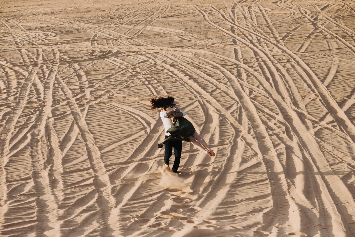 Couple embracing and spinning through warm desert sand dunes with textured tracks, cinematic adventure session