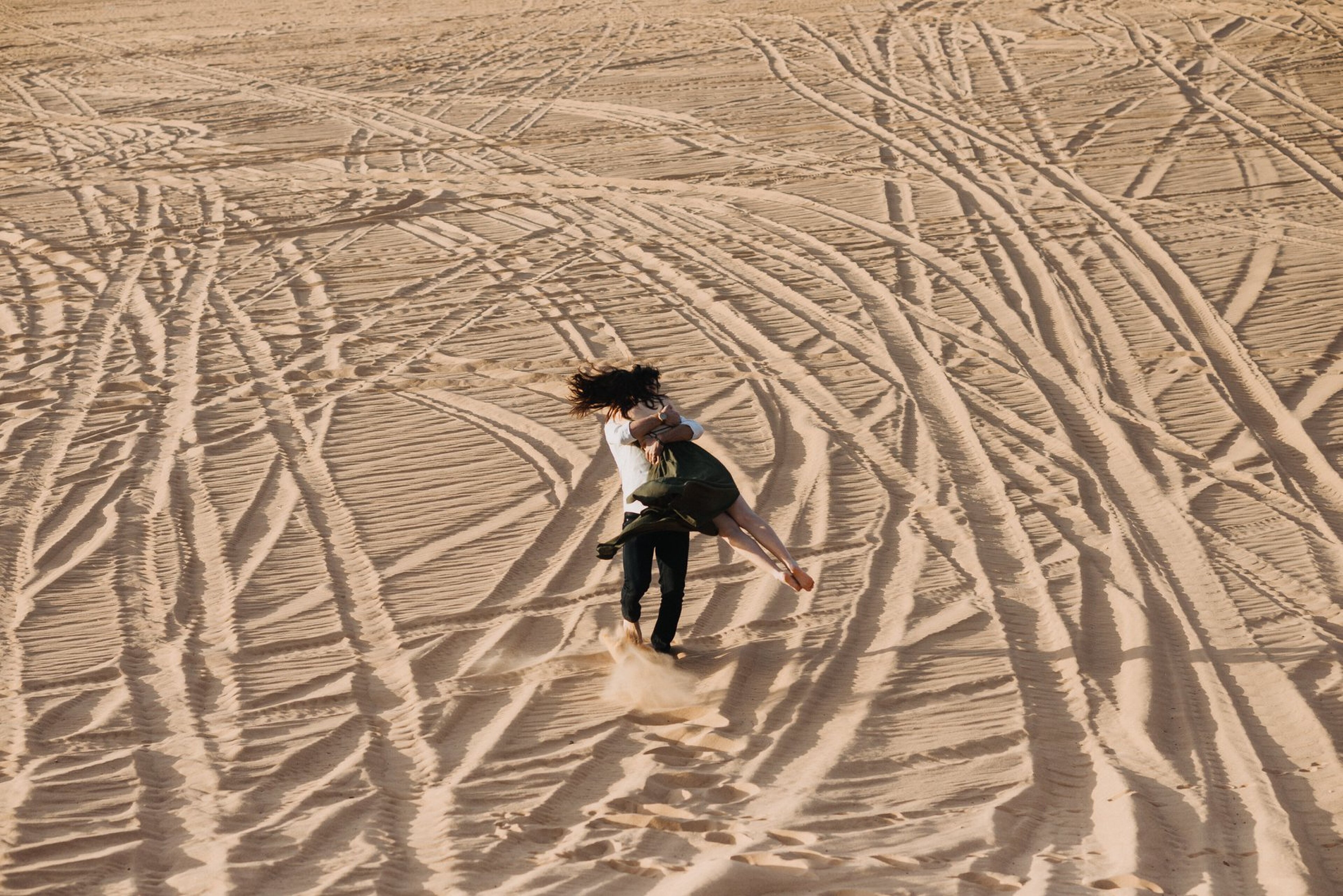 Couple embracing and spinning through warm desert sand dunes with textured tracks, cinematic adventure session